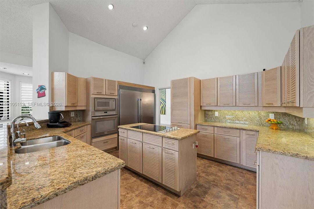 Kitchen featuring high vaulted ceilings, light wood-finish cabinetry, granite countertops, a stainless steel refrigerator, and a kitchen island with cooktop