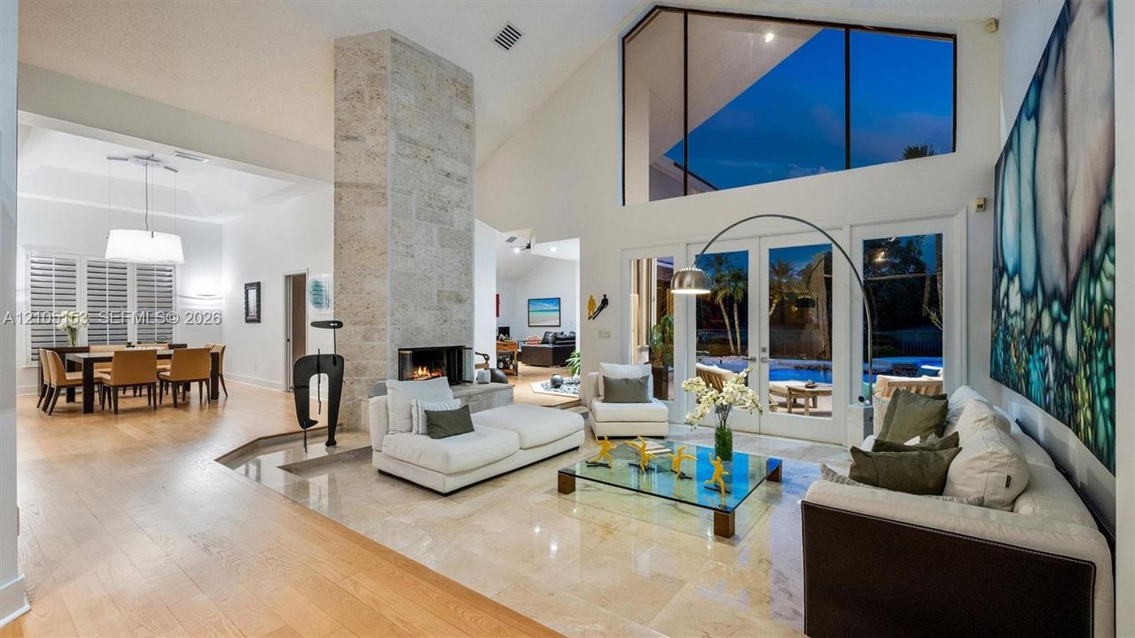 Soaring ceilings with transom windows, stone fireplace, light-toned tile flooring, and French doors opening to an exterior area with a pool