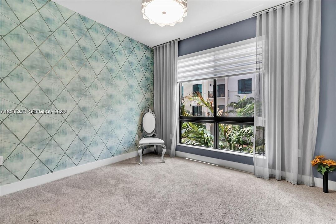 Room featuring geometric patterned accent wallpaper, large picture window with adjustable shades, neutral carpeting, and a semi-flush mount light fixture