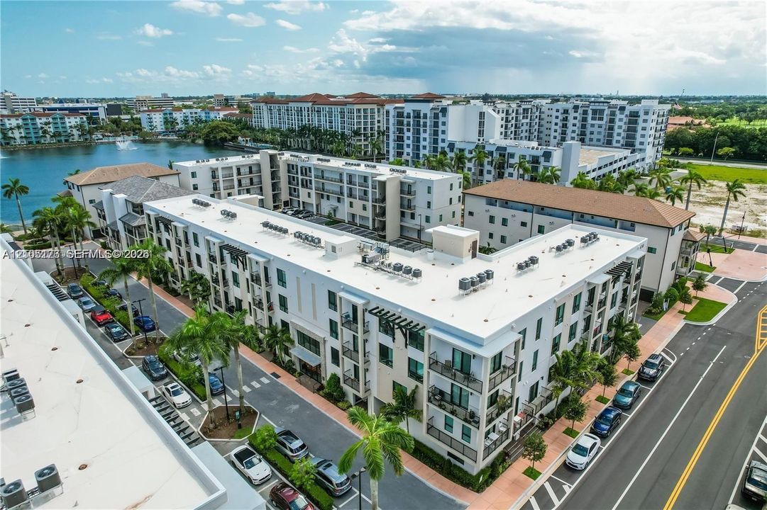 Aerial perspective of a multi-story residential building featuring a flat roofline, balcony structures, and integrated parking areas, situated near a water feature with paved walkways and palm trees