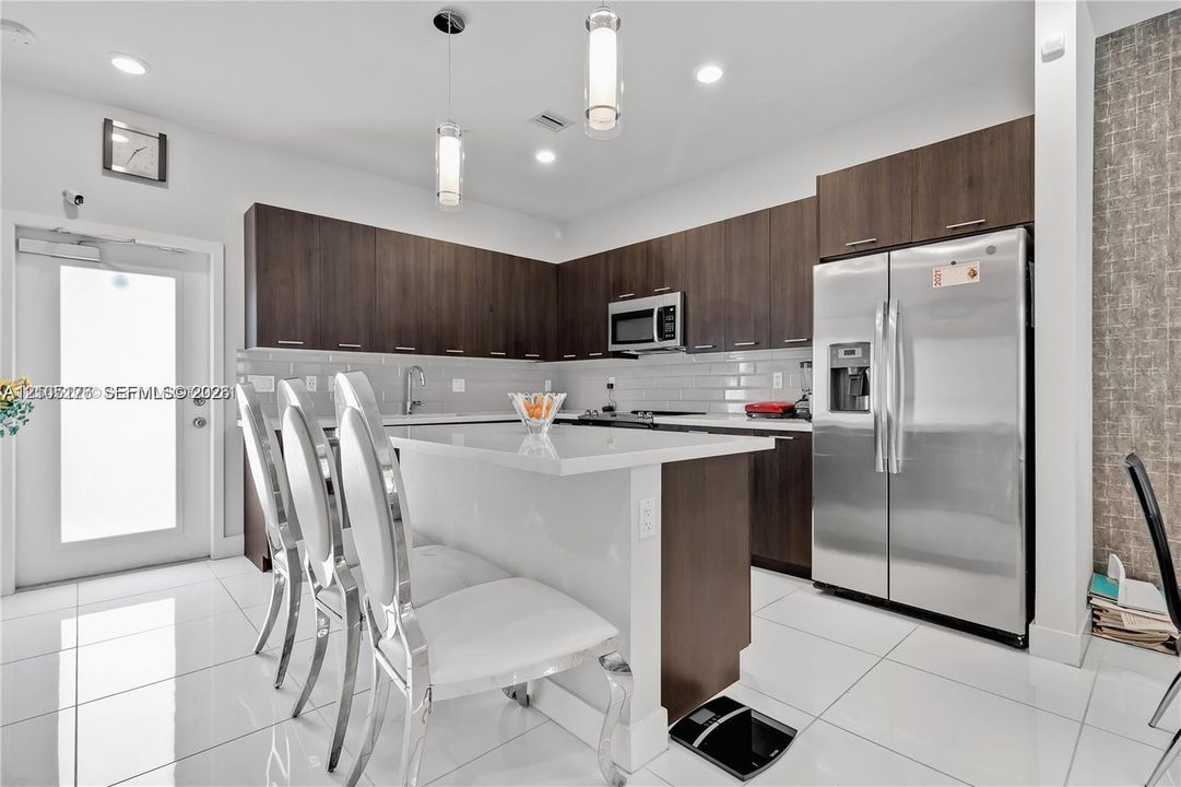 Modern kitchen featuring dark wood-finish cabinetry, a central island with white countertop seating, white tile flooring, stainless steel appliances, and minimalist pendant lighting