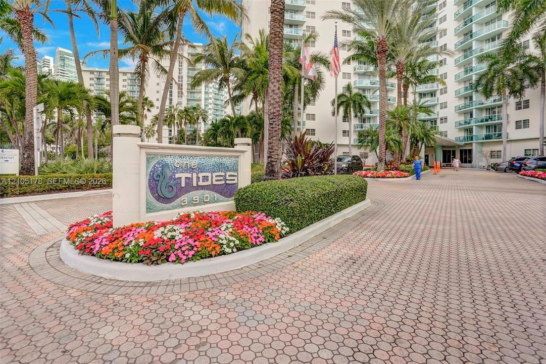 Paver driveway entrance featuring mature palm trees, manicured hedges, and floral landscaping surrounding the property monument sign