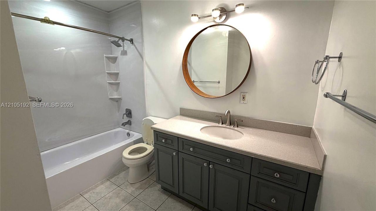 Bathtub with integrated shelving, gray vanity with light countertop, circular mirror with wood-finish frame, and tile flooring