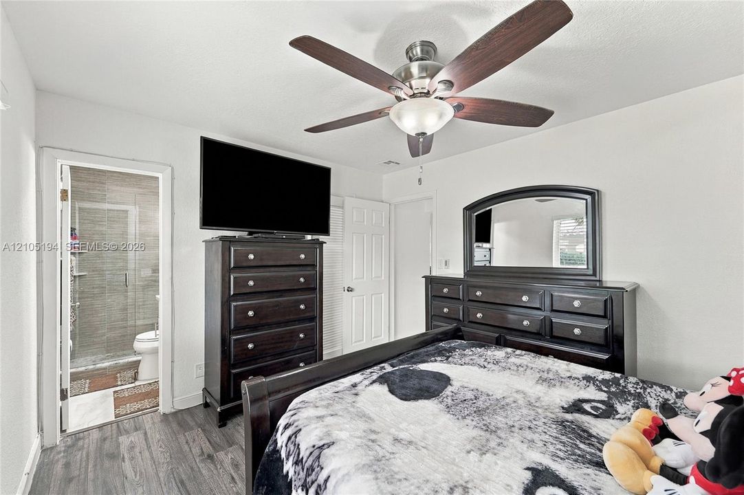 Room featuring wood-finish flooring, a multi-blade ceiling fan with integrated lighting, and a doorway entry to a tiled shower area