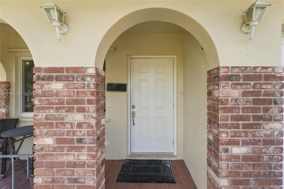 Covered entryway featuring arched architectural detailing, brick masonry columns, tile flooring, and wall-mounted exterior lighting fixtures