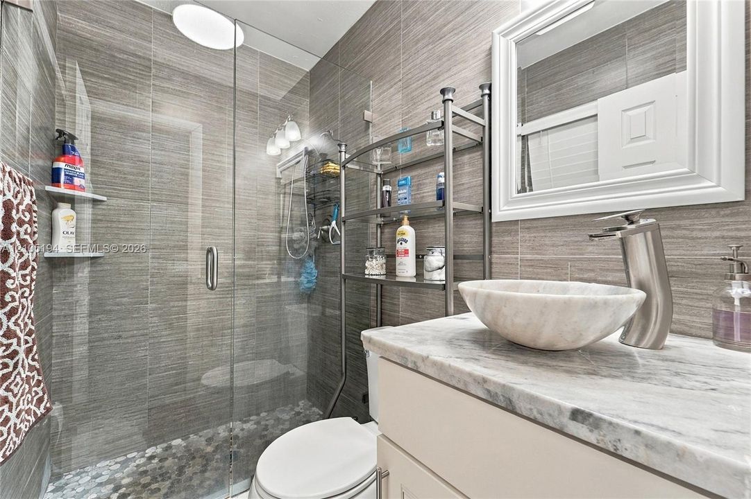 Bathroom featuring a vessel sink with a brushed-metal faucet, stone-finish countertop, frameless glass shower enclosure, pebble-tile shower flooring, and horizontal-grain wall tiling