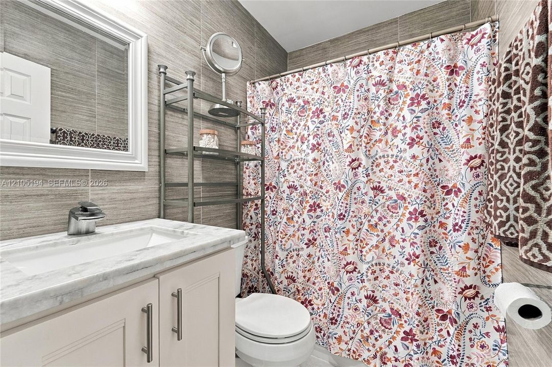 Bathroom featuring floor-to-ceiling textured wall tiling, vanity with a stone-finish countertop, undermount sink, and white cabinetry with brushed metal hardware