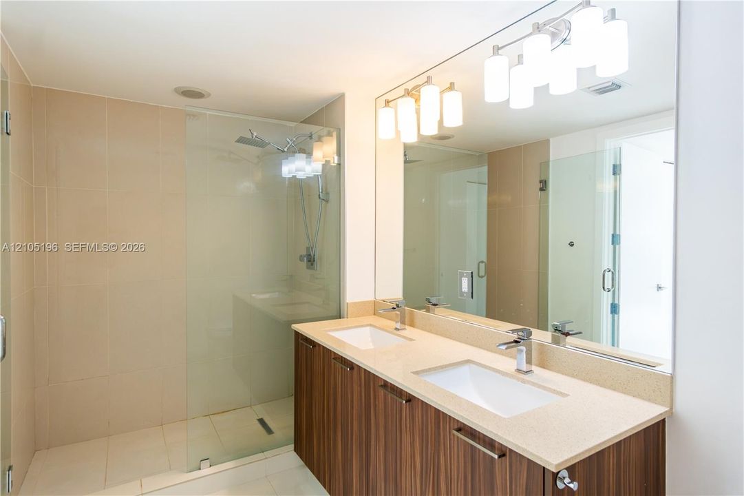 Dual vanity with stone countertop and integrated sinks, extensive mirror, and glass-enclosed shower with tiled walls
