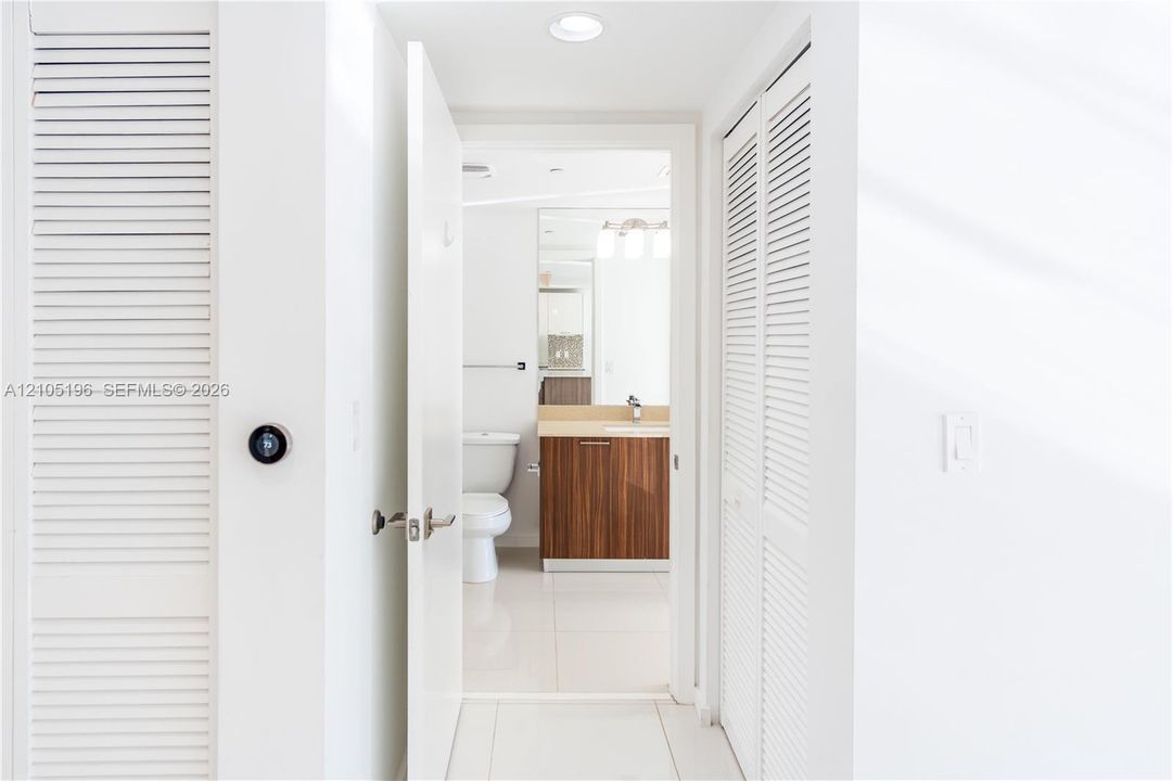 Hallway featuring white louvered doors and a smart thermostat, leading to a bathroom with a wood-finish vanity, light-toned countertop, and tile flooring