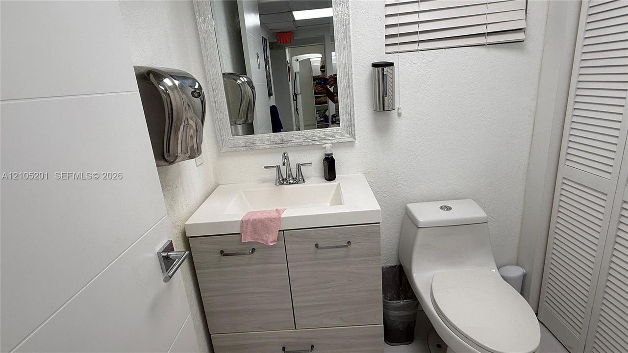 White vanity with wood-finish cabinetry and chrome fixtures, wall-mounted mirror, white wall-mounted toilet, and white louvered bifold doors