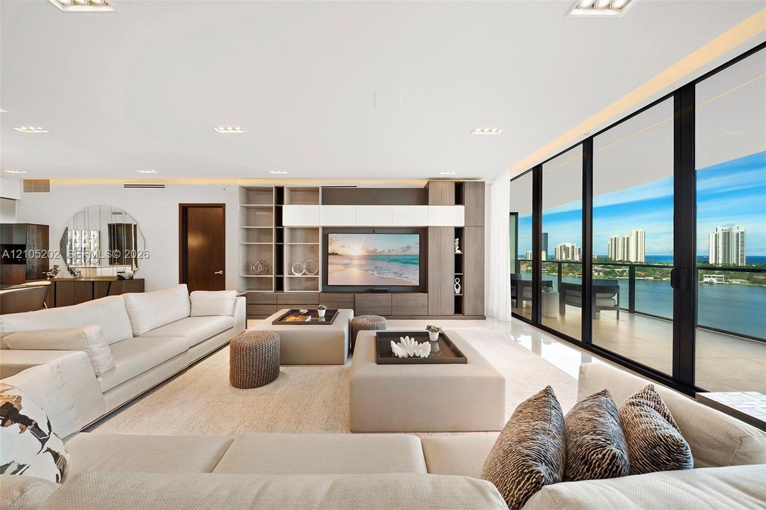 Floor-to-ceiling black-framed windows, built-in wooden media cabinetry, recessed ceiling lighting, glossy polished flooring, and expansive water backdrop