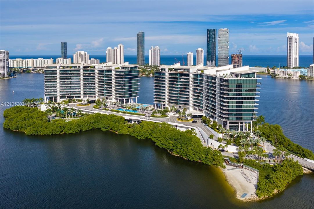 Waterfront residential complex featuring multiple glass-facade towers, deep wraparound balconies, an outdoor swimming pool, and tropical landscaping situated on a private peninsula