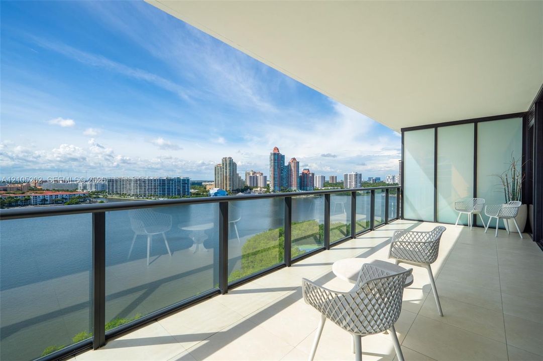 Covered balcony featuring oversized tile flooring, a glass panel railing with a dark metal frame, and water frontage
