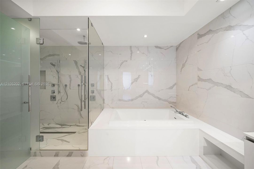 Enclosed glass shower featuring dual shower heads and a built-in recessed niche, complemented by a deep soaking tub surrounded by full-height stone-finish wall tiling and matching floor tile