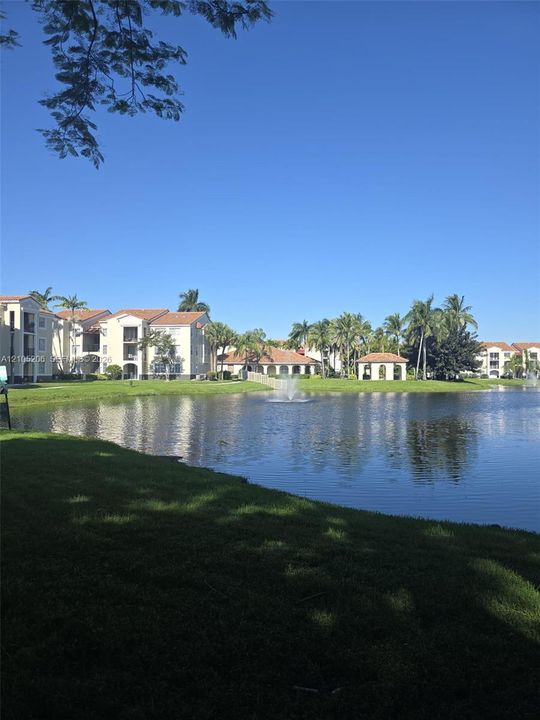 Community grounds featuring a central lake with a decorative fountain, manicured lawn, perimeter landscaping, and a covered pavilion