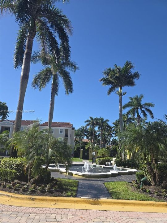 Multi-story building exterior featuring a decorative water fountain, paver stone hardscaping, mature palm trees, and manicured landscaping