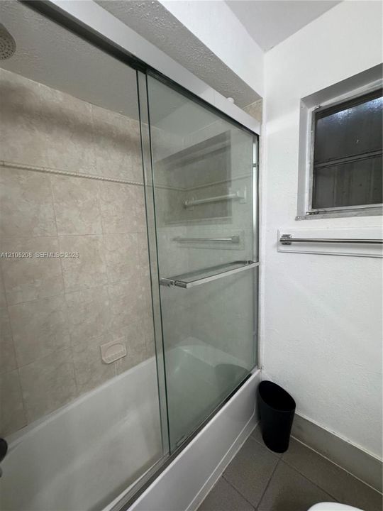 Bathtub with a glass sliding shower enclosure, light-toned wall tiling, and a window