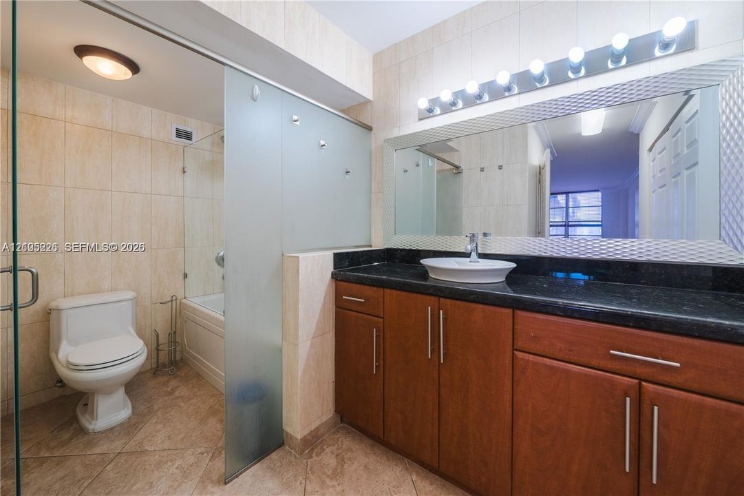 Bathroom featuring a dark stone-finish countertop, wood-finish cabinetry, vessel sink, full-width mirror with integrated lighting, and a glass-enclosed shower and tub combination