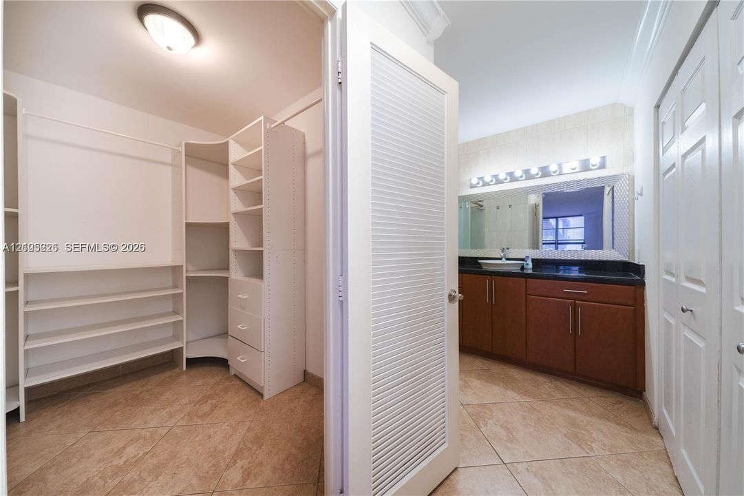 Walk-in closet featuring white built-in shelving, vanity with dark countertop and vessel sink, large wall-mounted mirror with integrated lighting, and tile flooring