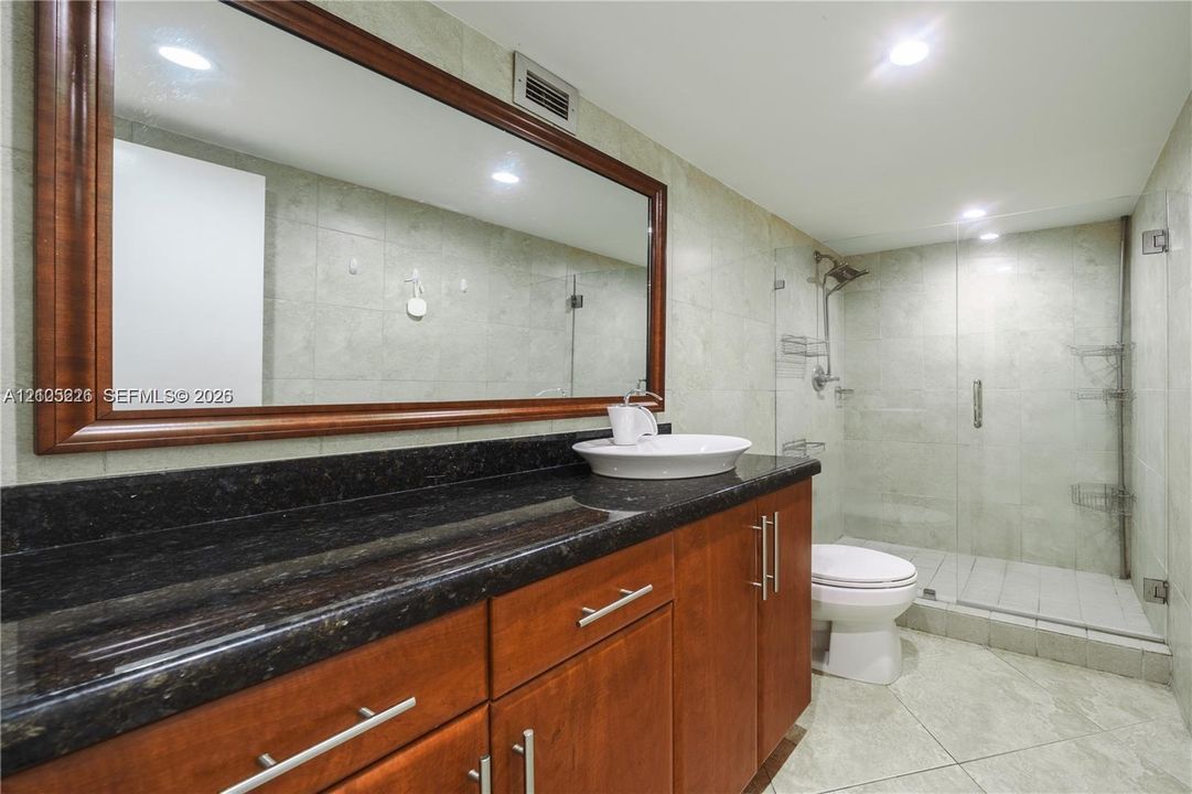 Wood-toned cabinetry featuring a dark stone countertop, a white vessel sink, a large wood-framed wall mirror, and a glass-enclosed shower with tile surround