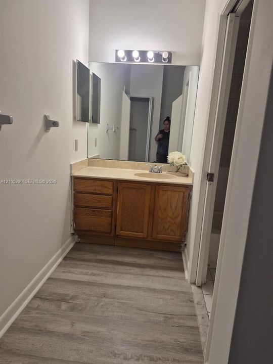 Vanity featuring wood-finish cabinetry, solid-surface countertop with an integrated oval sink, full-width wall mirror, and four-bulb light fixture, complemented by wood-finish flooring