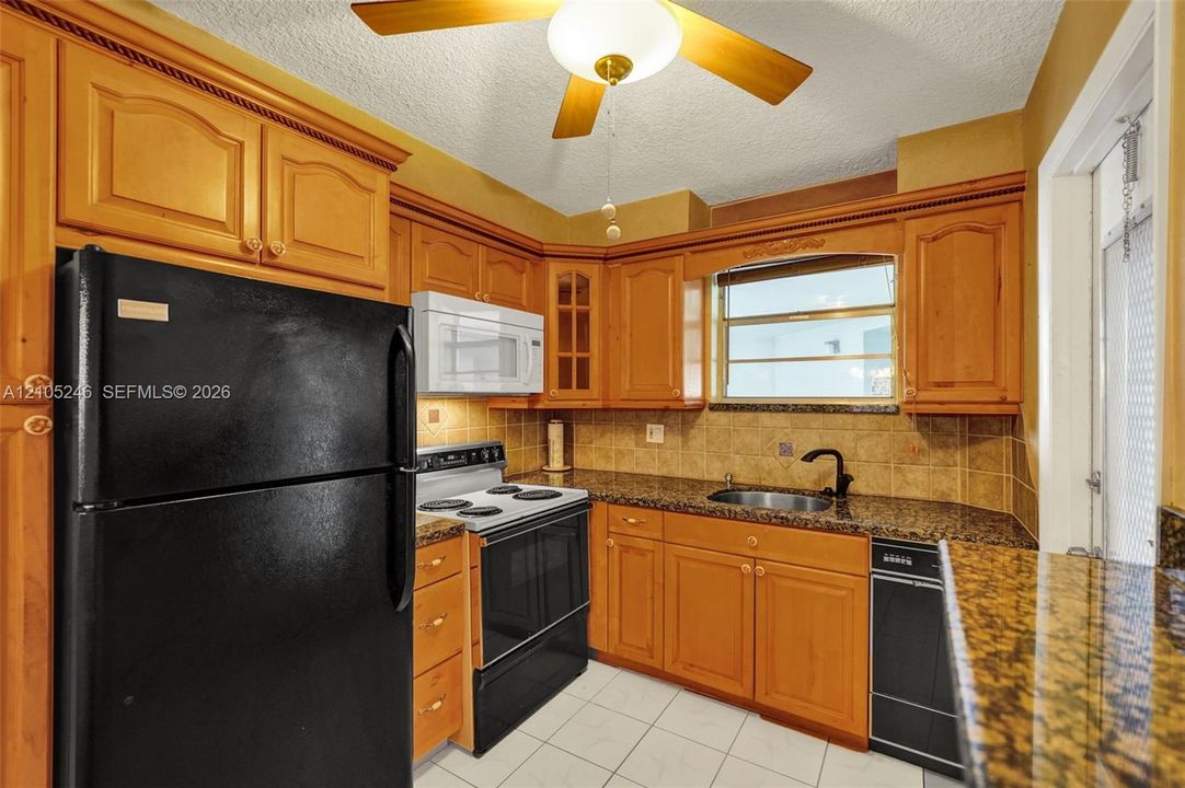 Kitchen featuring wood cabinetry, stone countertops, a tile backsplash, tile flooring, and integrated black appliances