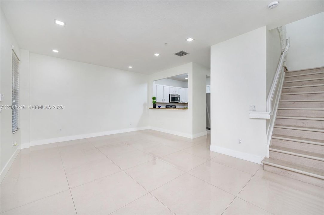 Open-concept living space featuring large format floor tiles, recessed ceiling lighting, a half wall with a countertop looking into the kitchen, and a staircase with a white railing