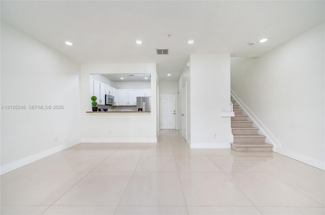 Spacious main living area featuring large format tile flooring, recessed lighting, a pass-through kitchen with white cabinetry, stainless steel appliances, and a staircase with a white railing