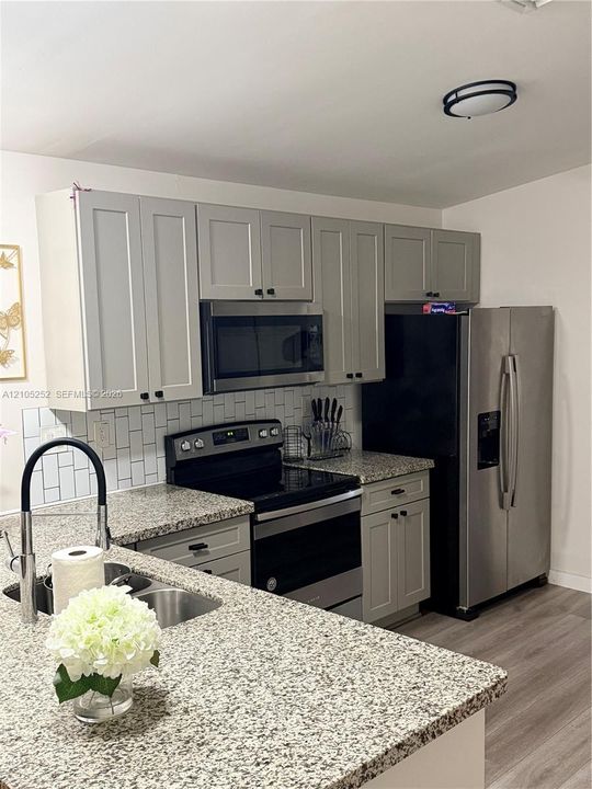 Updated kitchen featuring granite countertops, stainless steel appliances, light gray shaker cabinetry, a subway tile backsplash, and wood-finish flooring
