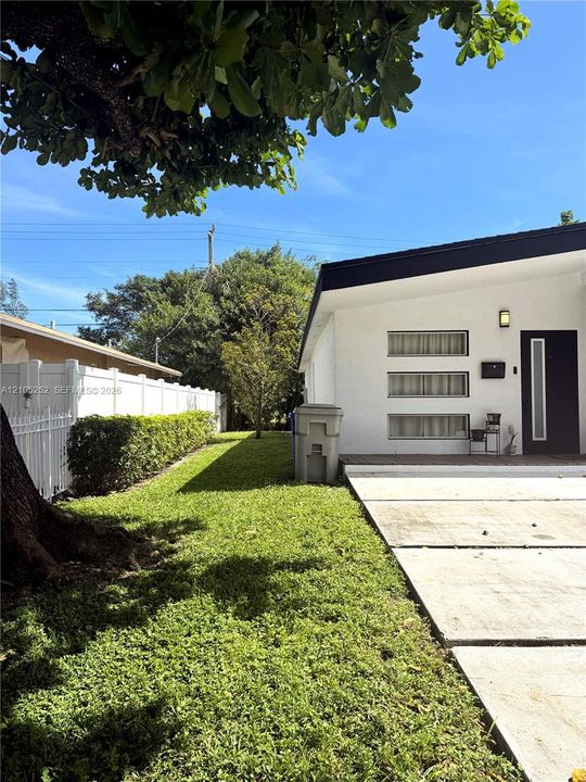 White exterior with black trim, concrete walkway, green lawn, white picket fence, and mature landscaping