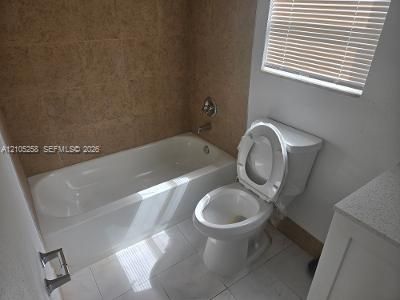 White bathtub with chrome fixtures, tiled shower surround, white toilet, white tile flooring, and a window with blinds