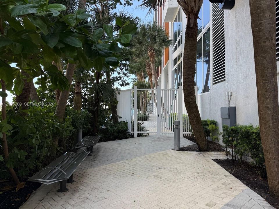 Exterior walkway featuring interlocking paver stones, white metal perimeter fencing, integrated metal benches, and mature palm trees