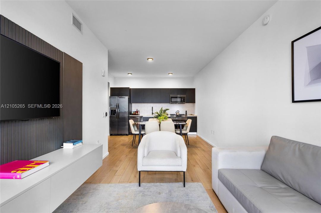 Modern interior featuring wood-finish flooring, a matte-finish accent wall with a wall-mounted media console, dark cabinetry, and white tile backsplash