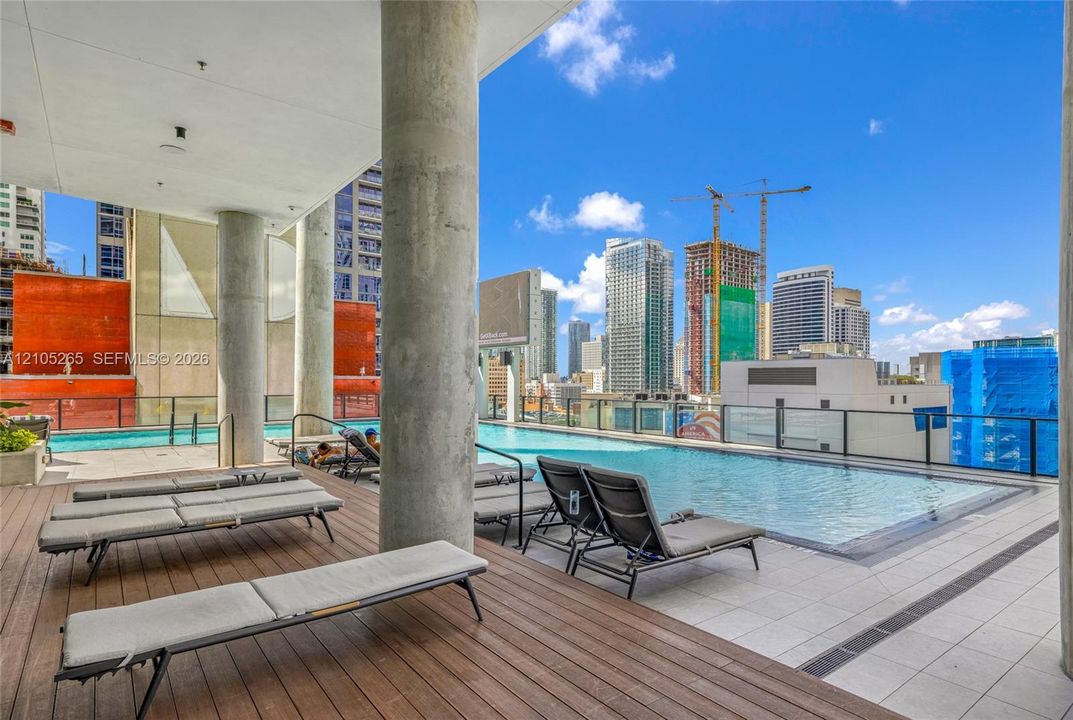 Rooftop swimming pool with tile surround, wood-finish deck, concrete structural columns, and glass panel railing