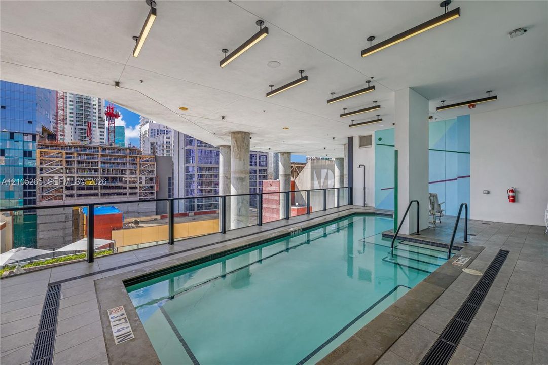 Covered swimming pool featuring linear overhead lighting, structured support columns, and stone-tile decking with integrated drainage