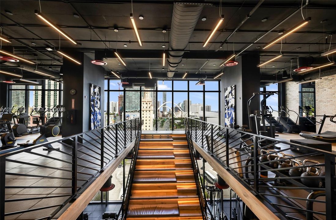 Industrial-style fitness center featuring an open-concept layout, floor-to-ceiling glass windows, exposed ductwork, wood-tread staircase with integrated lighting, and matte black metal railings