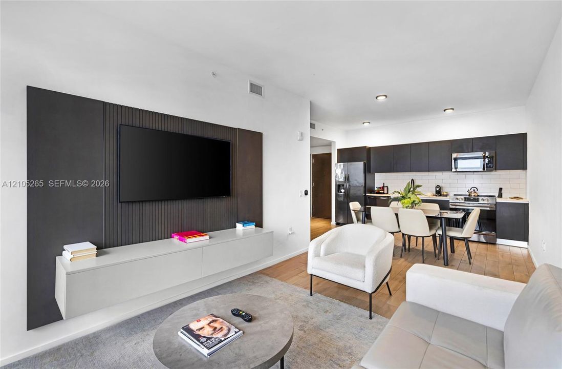 Living area featuring a custom dark-toned ribbed media wall with a built-in light gray floating console, complemented by wood-finish flooring and kitchen cabinetry finished in a dark stain with a subway tile backsplash
