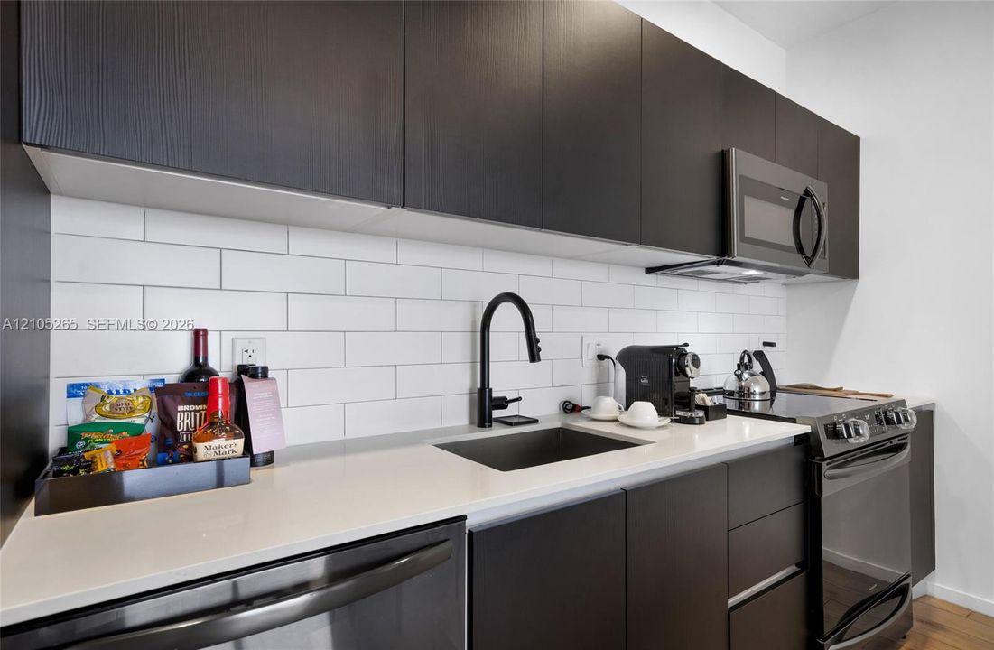 Modern kitchen featuring dark slab-style cabinetry, light-toned countertops, white subway tile backsplash, matte black hardware, and stainless steel appliances