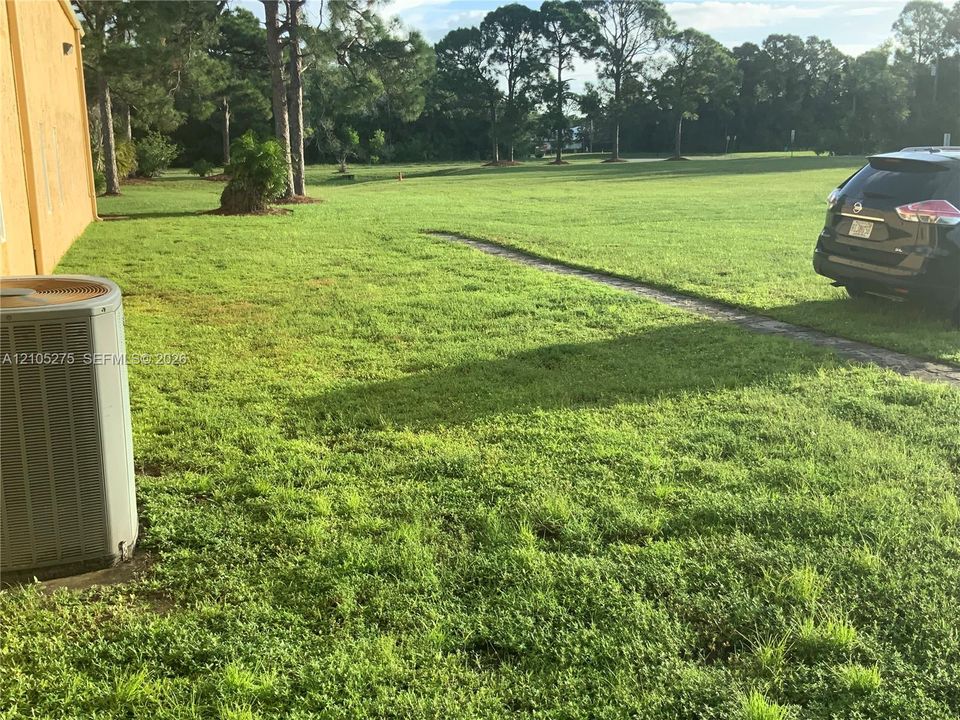 Expansive manicured lawn with mature tree-lined backdrop, HVAC unit, and circular driveway with vehicle parking