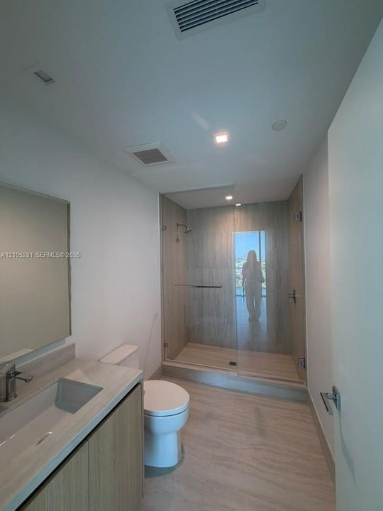 Bathroom featuring a wood-finish vanity with a neutral stone countertop, frameless glass shower enclosure with floor-to-ceiling tile, and integrated recessed lighting