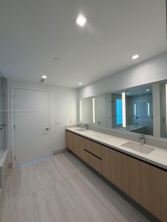 Double vanity featuring light-toned wood cabinetry, dual undermount sinks, stone-finish countertops, wall-to-wall integrated mirror with vertical LED lighting, and neutral large-format tile flooring