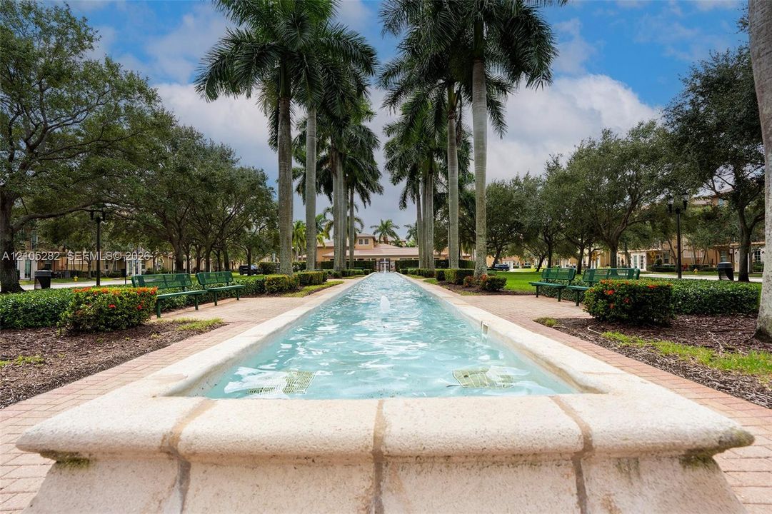 Linear water feature with stone coping, lined by mature palm trees, manicured hedges, and paver pathways