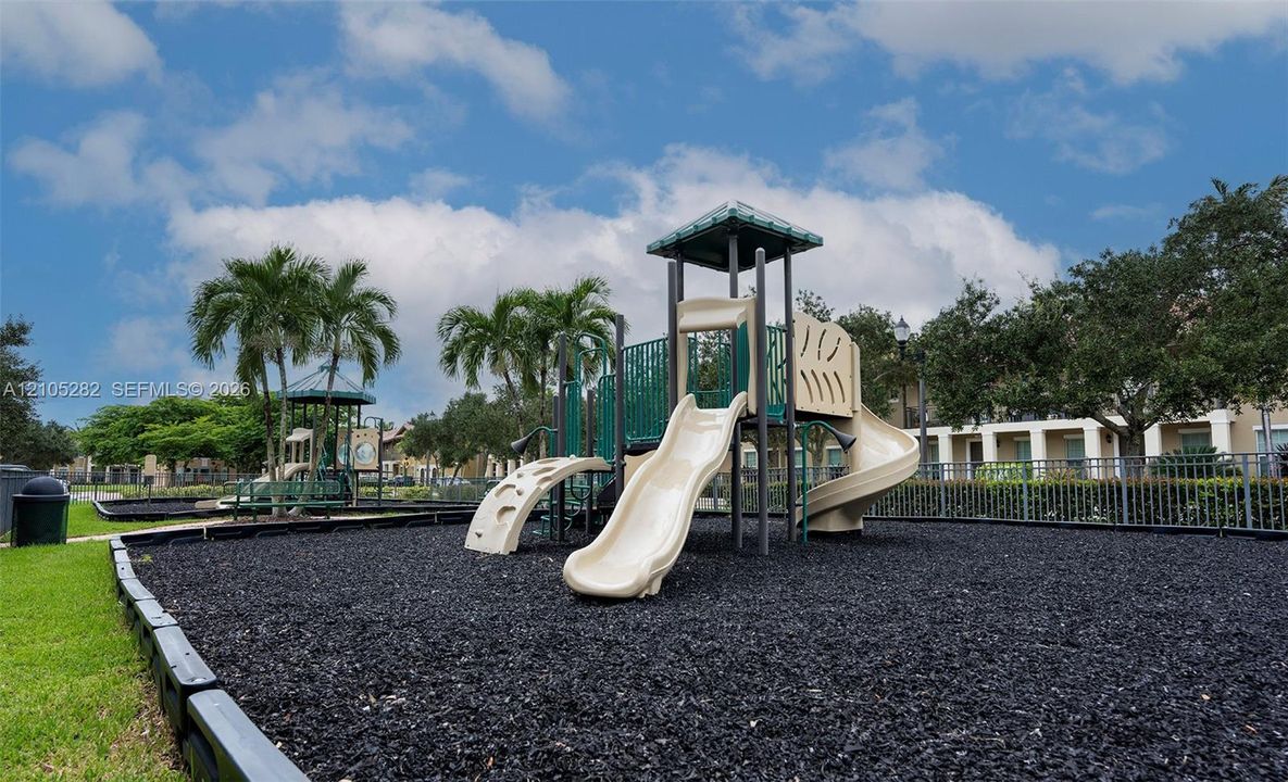 Recreational area featuring a multi-level climbing structure with slides, black rubber mulch surfacing, and metal perimeter fencing