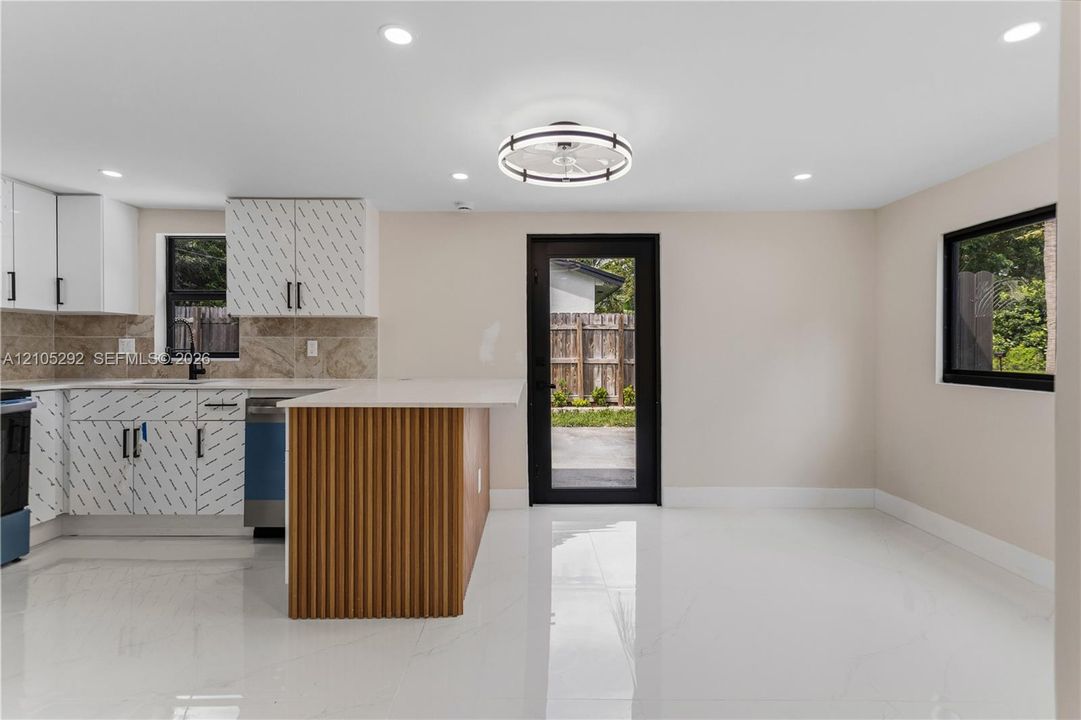 Modern kitchen featuring white cabinetry, stone-finish backsplash, built-in dishwasher, and a center island with a wood-slat base and light-toned countertop