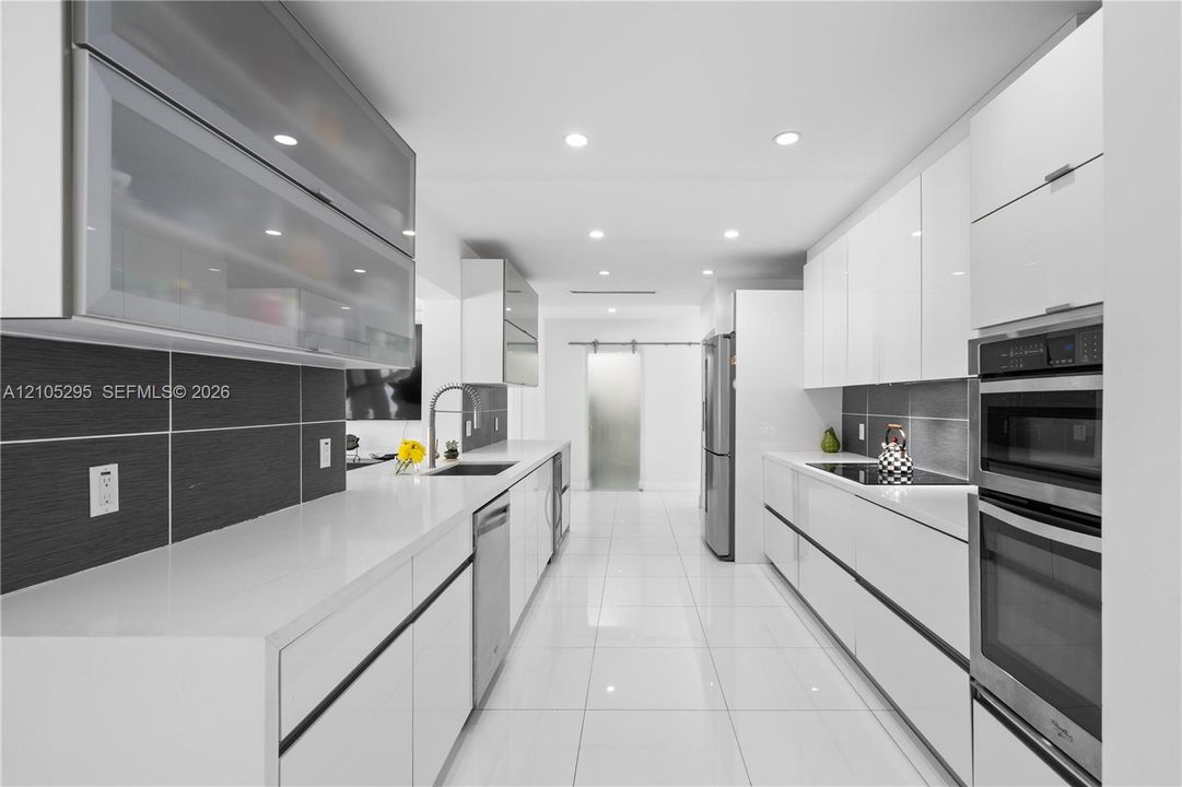 Modern kitchen featuring high-gloss white cabinetry, expansive stone countertops, stainless steel appliances including a double wall oven, dark linear tile backsplash, and glass polished tile flooring