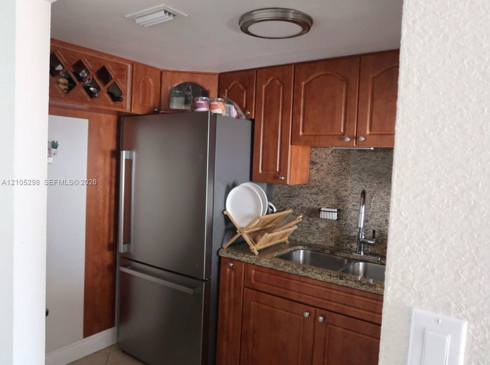 Kitchen featuring wood cabinetry, stainless steel refrigerator, double basin sink with a gooseneck faucet, and stone-finish countertops with a matching backsplash