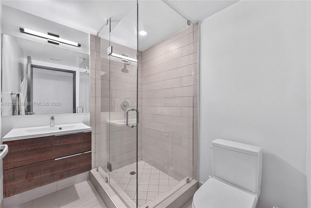 Floating wood-finish vanity with an integrated sink, large vanity mirror, glass-enclosed shower with tile surround, and an elongated toilet