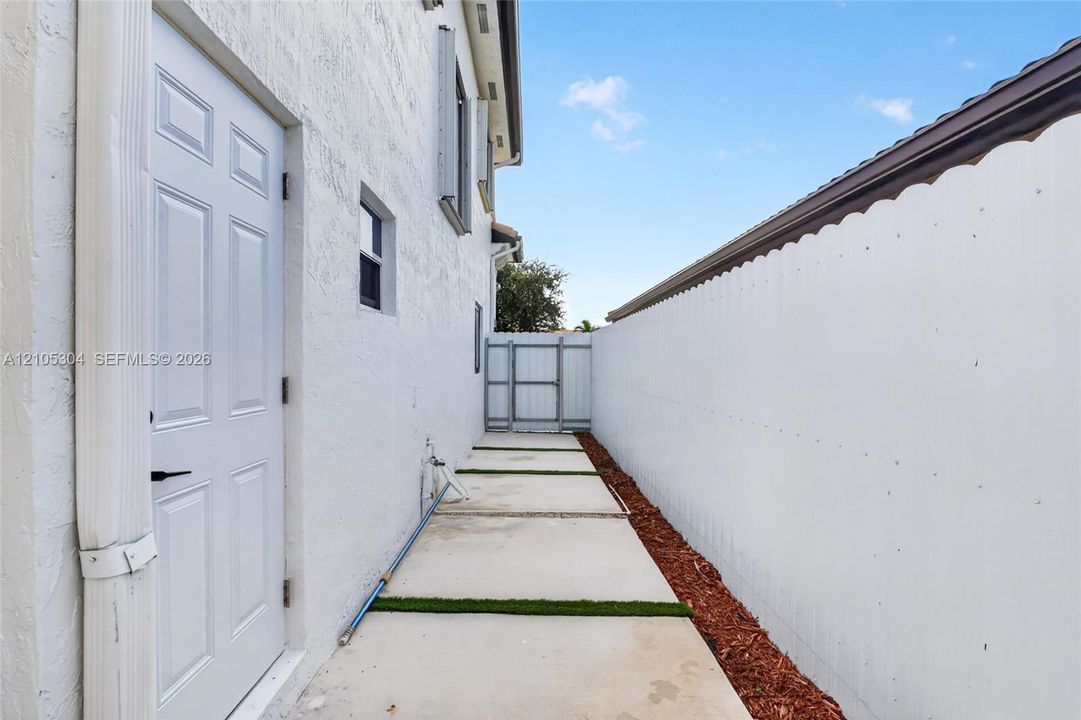 Side exterior featuring a stucco facade, concrete paver path with grass inlay, mulched landscape border, solid perimeter wall, and gated entry