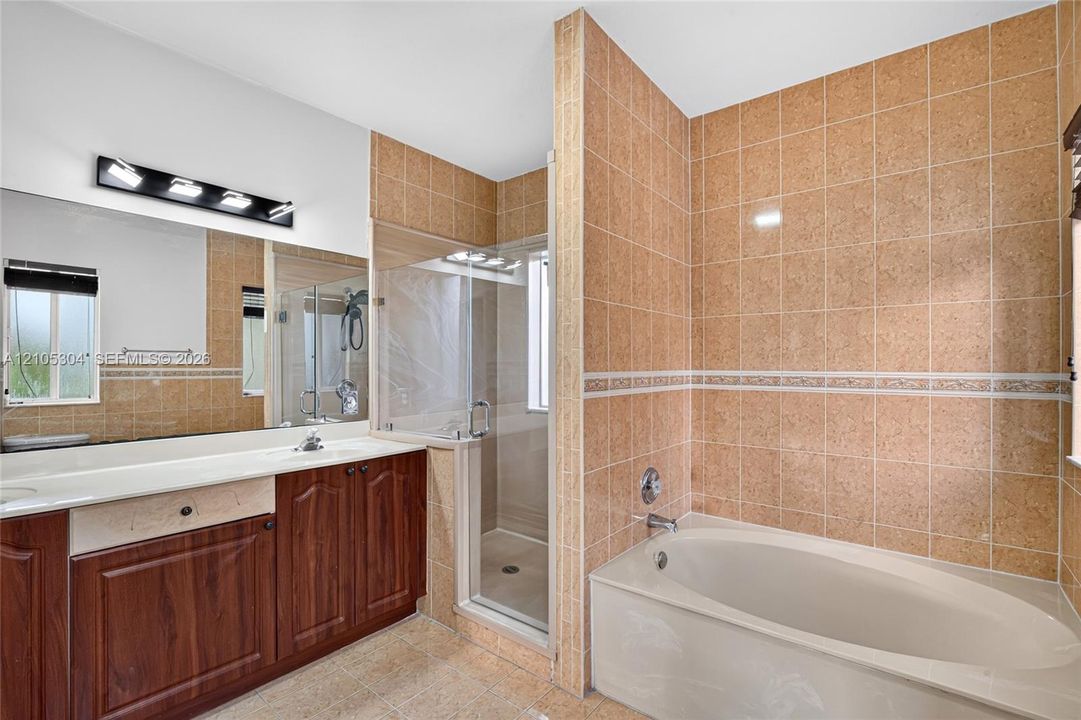Bathroom featuring a wood-finish vanity with a light-toned countertop, a soaking tub with tile surround, a glass-enclosed shower, a large wall-mounted mirror, and vanity lighting