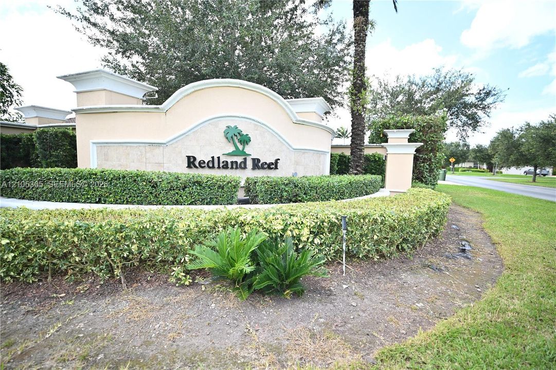 Redland Reef community entrance featuring a stucco and stone-finish monument sign, lush green landscaping, and mature foliage